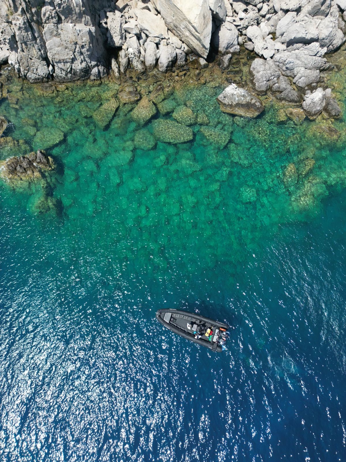 The RIB at anchor on the edge of turquoise shallows beside a rocky shore