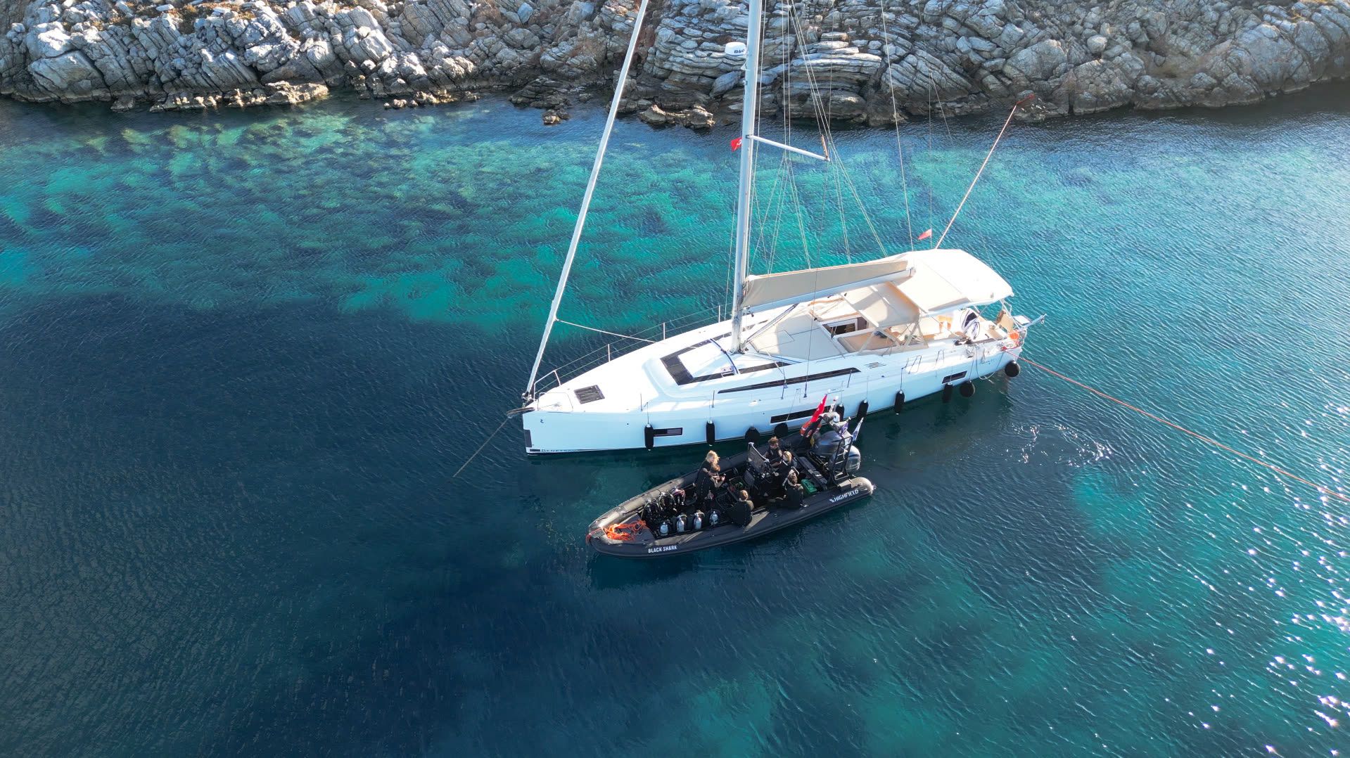 The DiveTalia liveaboard anchored in a rocky cove, the Highfield RIB alongside with divers and tanks aboard