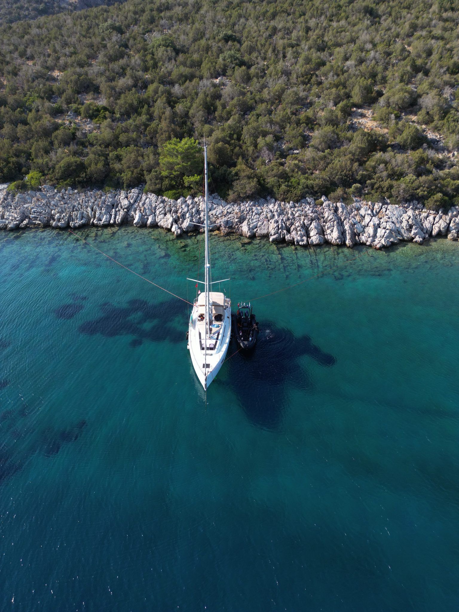 The yacht and RIB seen straight down over turquoise water by a rocky shore