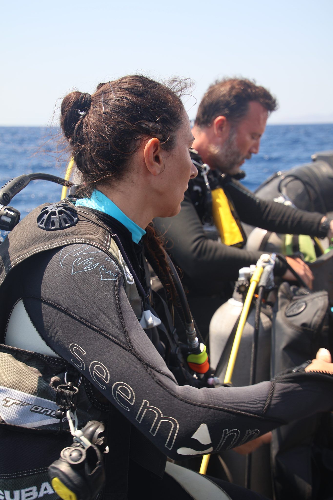 A diver gearing up in profile, another out of focus behind her, open sea beyond