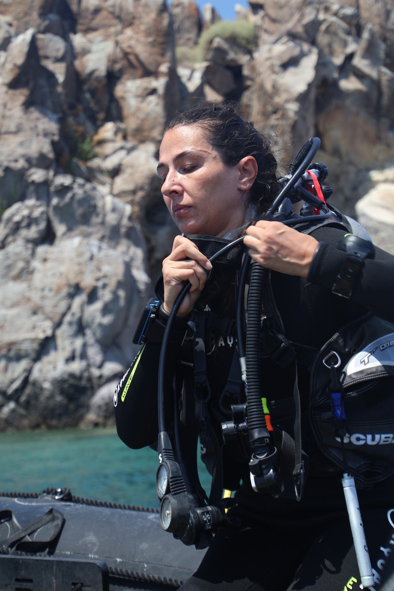 A diver fastening her hood, a rocky cliff and turquoise water behind her