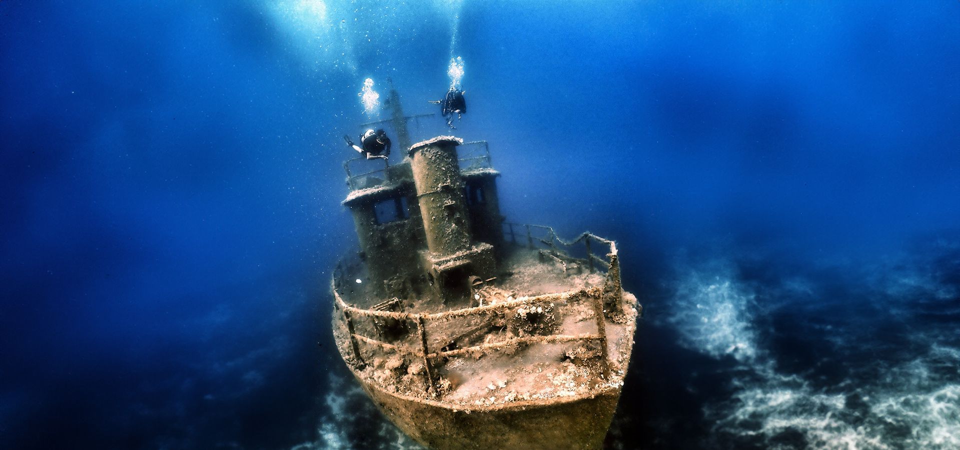Two divers above the bow of a wreck standing upright on the seabed, its mast and rails rising into deep blue water