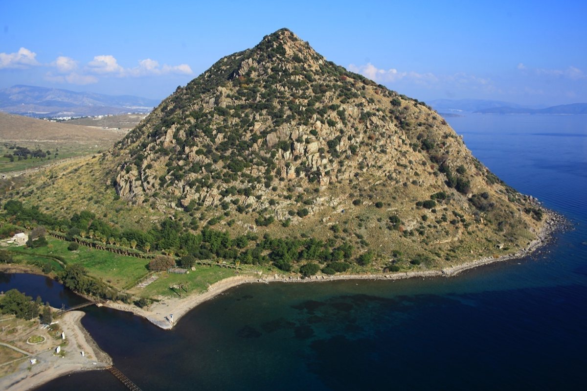 The conical hill above Aspat Bay seen from the air, rising straight out of the sea, with the beach, the jetty and the anchorage below it and the Aegean stretching beyond.