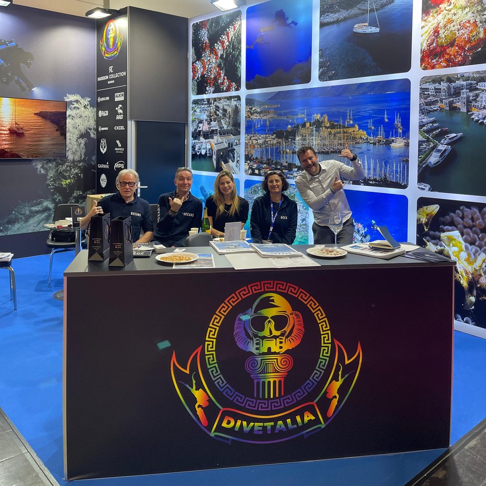 The DiveTalia team behind the counter of their stand at boot Düsseldorf, the crest across the front of the counter, a wall of Bodrum and underwater photographs behind them and partner logos on the side panel.