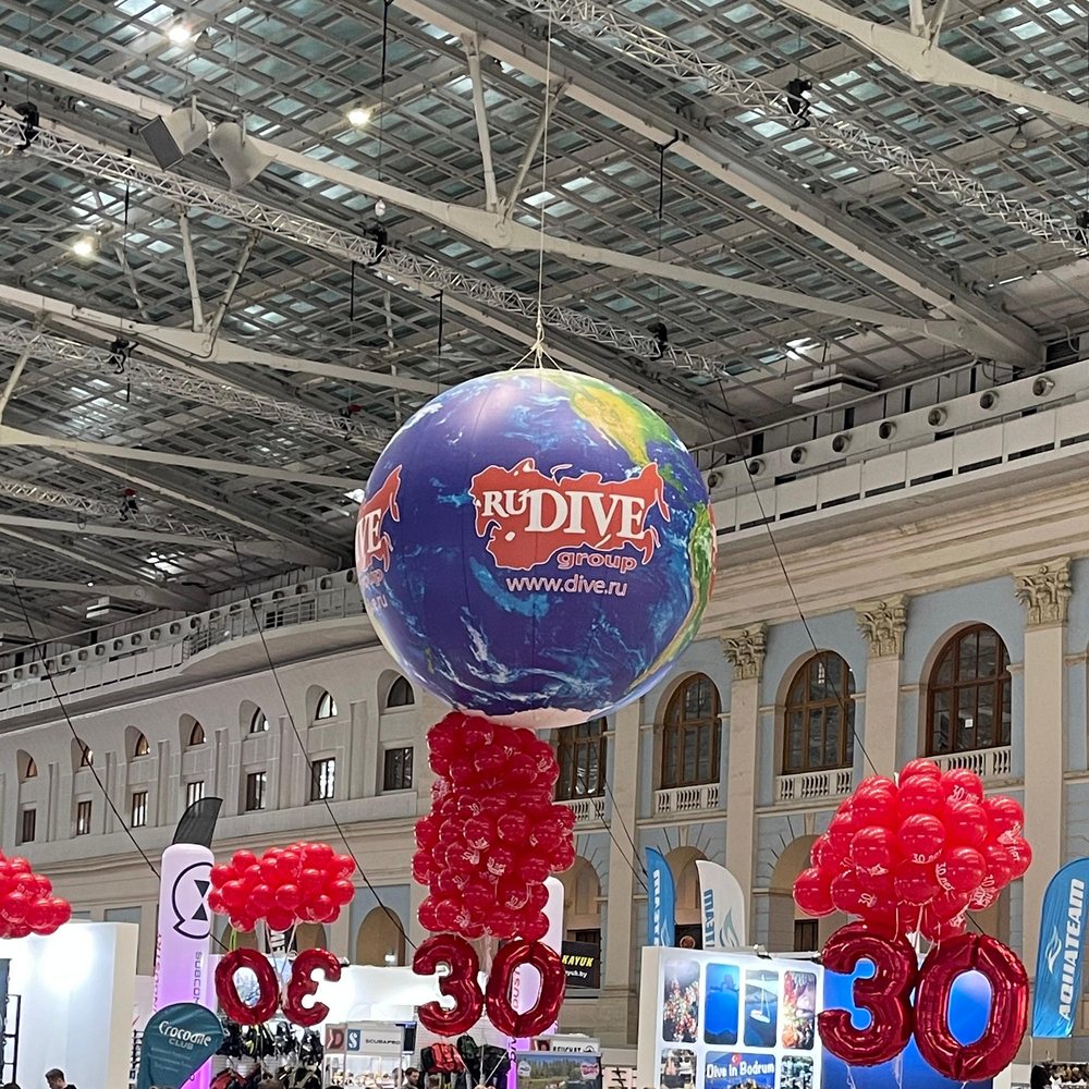 A large globe balloon hanging above the Moscow Dive Show floor in Gostiny Dvor, with clusters of red balloons and numeral balloons over the stands below.
