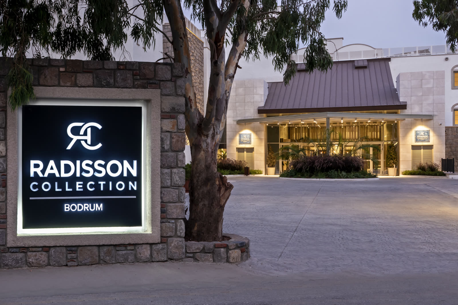 The lit Radisson Collection Bodrum entrance at dusk, the hotel sign glowing beside a eucalyptus tree and the canopied doors beyond